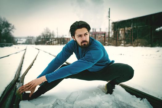 Active young man crouching and doing exercises in the public place among old railroad during the winter training outside in. Copy space.