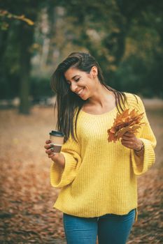 Cute young woman enjoying in sunny forest in autumn colors. She is holding golden yellow leaves and cup of cofee.