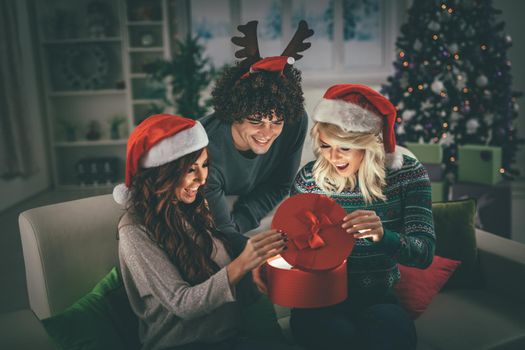 Cheerful beautiful young friends enjoying Christmas at home, opening gift and surprisingly looking what is inside.