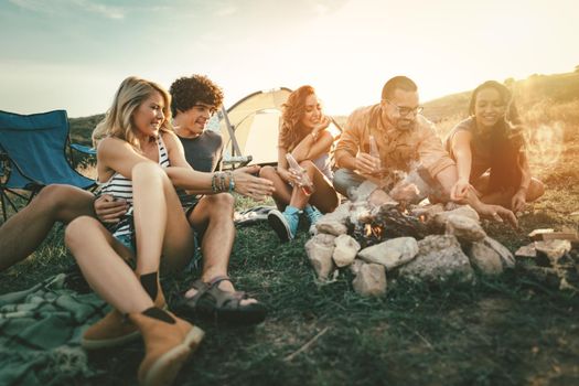 Happy young friends enjoy a sunny day at the mountain. They're laughing and preparing campfire near tent.