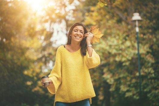 Cute young woman enjoying in sunny forest in autumn colors. She is holding golden yellow leaves and cup of cofee.