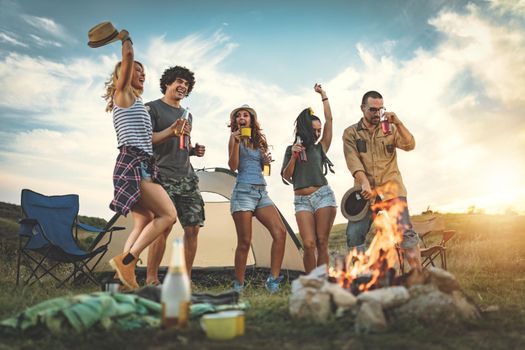 Happy young friends enjoy a nice day in nature. They're holding beer bottles, laughing, dancing, and talking happy to be together.