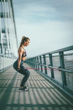 Muscular young woman is doing strong fit body training with elastic rubber band on the bridge.