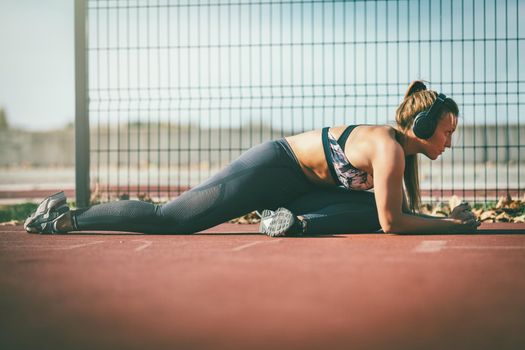 Young female runner, with headphones on her ears, doing stretching exercise and preparing for morning workout.