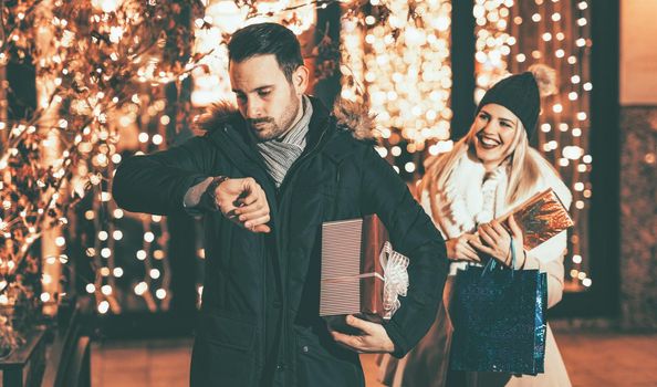 Young man worried looking at the watch in a Christmas night and waiting his girlfriend an appointment. She is late, and surprised stand behind him.