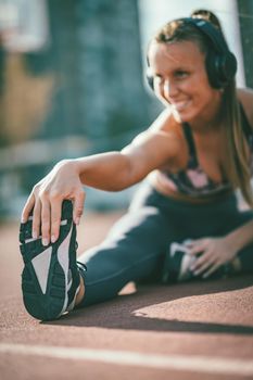 Young female runner, with earphones on her ears, doing stretching exercise on a public place, preparing for morning workout.