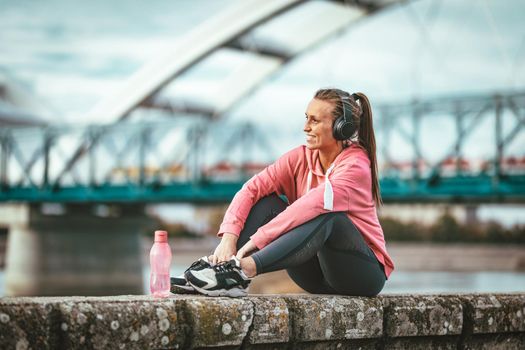 Young fitness woman with headphones is resting after hard training on the wall by the river bridge with bottle of a water.