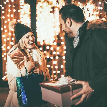 Young beautiful cheerful couple celebrating Christmas in the city street and giving many gift to each other. 