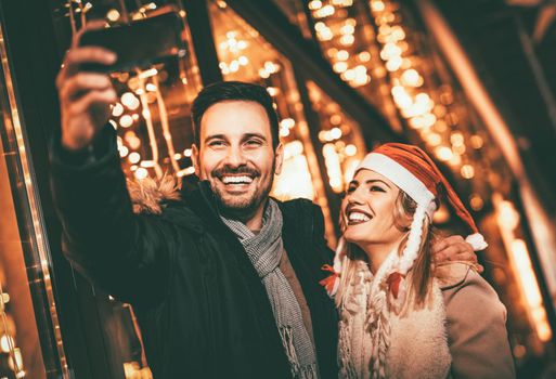 Young beautiful cheerful couple taking a selfie in the city street at new year's night with a lot of lights on background. 