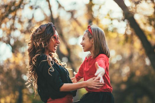 Beautiful young mother and her happy daughter having fun in the forest in sunset. They are hugging, smiling and looking each other.