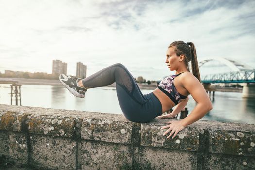 Focused young sport woman doing sit-up exercises on the wall during outdoor cross training by the river.