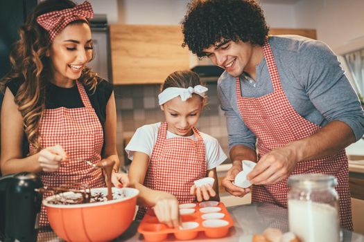 Happy parents and their daughter are preparing cookies together in the kitchen. Little girl helps to her parents to put paper cups into molds.