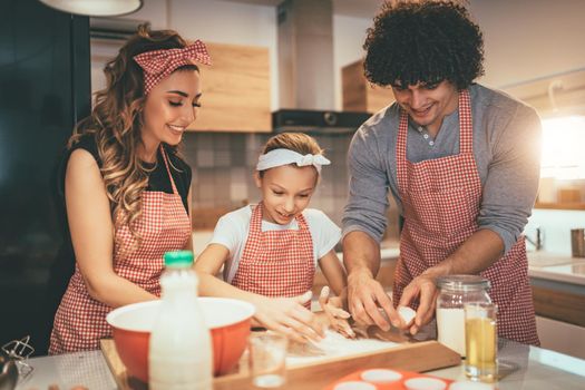 Happy parents and their daughter are preparing dough for cookies together in the kitchen. Little girl helps to her parents to mix flour with eggs.