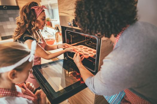 Happy parents and their daughter put cookies in the oven to bake, making fun.