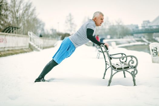 Active senior man athlete doing push ups on the bench and doing exercises by the river during the winter training outside in. Copy space.