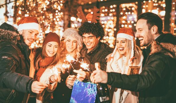 Cheerful six young friends having fun in city mall in Christmas night, while using sparklers, with bright shop window in background.