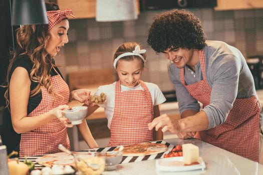 Happy parents and their daughter are preparing meal together in the kitchen. Little girl and her father are putting ketchup and origano on the pizza.