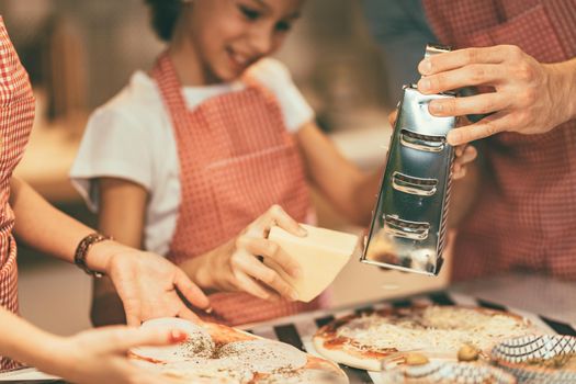 Happy parents and their daughter are preparing meal together in the kitchen. Little girl and her father are putting cheese on the pizza.