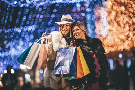 Young beautiful cheerful female friend with colorful shopping bags having fun and walking in the city street at night at Christmas time.