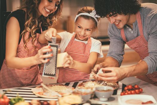 Happy parents and their daughter are preparing meal together in the kitchen. Little girl and her mother are putting cheese on the pizza and her father is cutting mushrooms.