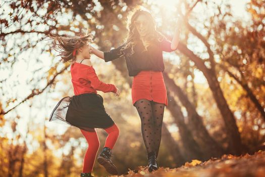 Beautiful young mother and her happy daughter having fun in the forest in sunset. They are holding hands, laughing and jumping.