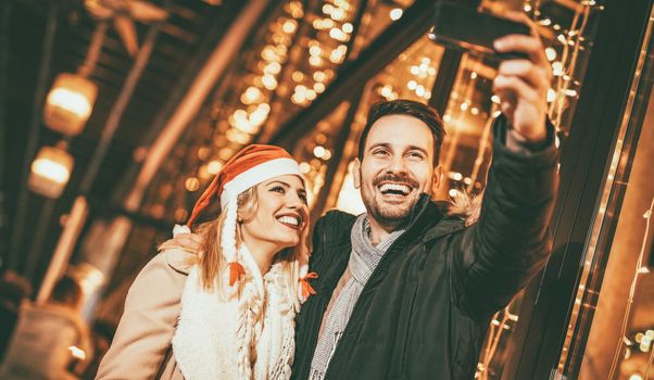 Two beautiful cheerful young people taking selfie in the shopping mall with bright shop window in background.   
