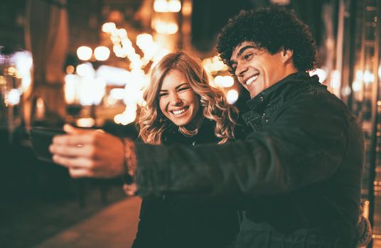 Young beautiful cheerful couple taking a selfie in the city street at new year's night with a lot of lights on background. 