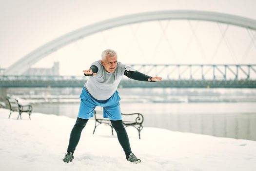 Active senior man stretching and doing exercises by the river during the winter training outside in. Copy space.