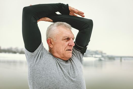 Active senior man stretching and doing exercises by the river during the winter training outside in.