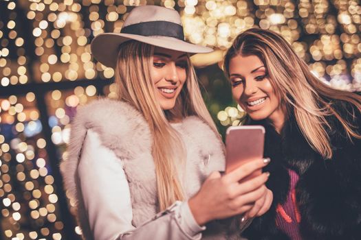 Cheerful young female friends are having fun in city street at the Christmas night. They are surfing on social network at smartphone and talking something funny.