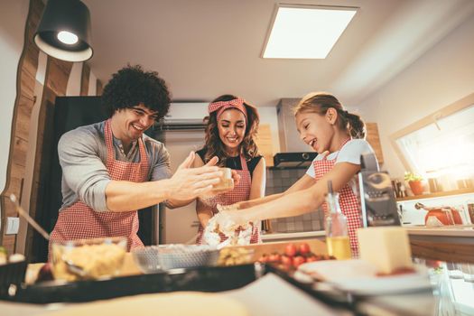 Happy parents and their daughter are preparing dough for pizza together in the kitchen. Little girl helps to her parents to mix dough on the table.