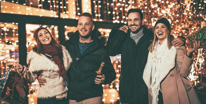 Two young cheerful couple having fun at the Christmas outdoor party in the city street at night and with a lot of lights on background.