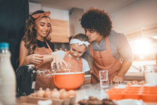 Happy parents and their daughter are preparing cookies together in the kitchen. Little girl helps to her parents pouring the oil in dish with dough.