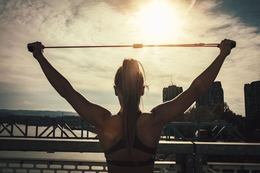 Rear view of a muscular young woman who is doing strong fit body training with elastic rubber band on the bridge in sunset.