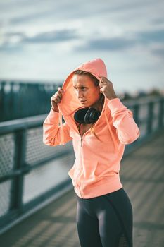 A young sport woman dresses up a sweatshirt and prepares for morning workout on a river bridge.