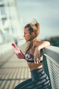 Young fitness woman is resting after hard training on the river bridge and drinking water.