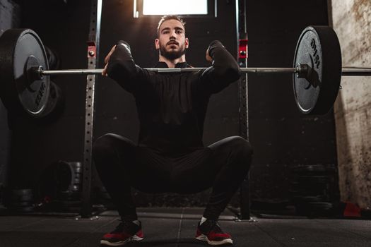 Young muscular handsome man is doing squat exercise with barbell at the gym. 