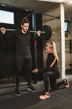 Young muscular man doing exercise with barbell at the gym, with support of his personal female trainer. 