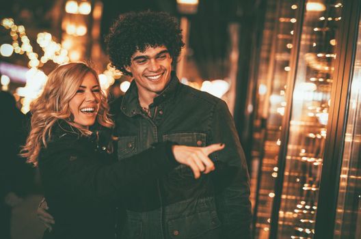 Young beautiful cheerful couple having fun in the city street at New Year time pointing at something through the window shop.