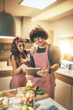 Young happy couple is enjoying and preparing healthy meal in their kitchen and reading recipes on the tablet.