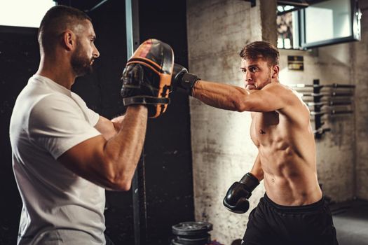 Young muscular man is practicing boxing exercise in the gym support of her personal trainer. 