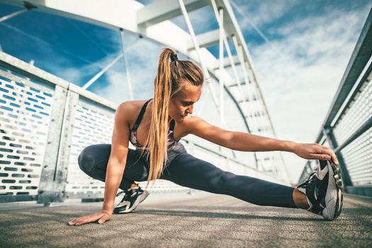 Young female runner doing stretching exercise on a river bridge, preparing for morning workout.