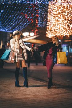 Young beautiful two cheerful sisters, with colorful shopping bags, having fun and walking in the night city street at Christmas time.