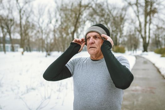Active senior making pause, puts a hat on the had, during jogging in public park during the winter training outside in. Copy space.