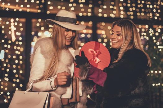 Young beautiful two cheerful friends are celebrating Christmas on the city street, giving presents to each other. 