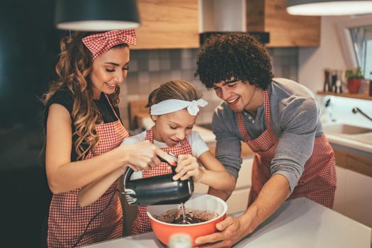 Happy parents and their daughter are preparing cookies together in the kitchen. Little girl helps to her parents mixing dough with mixer.