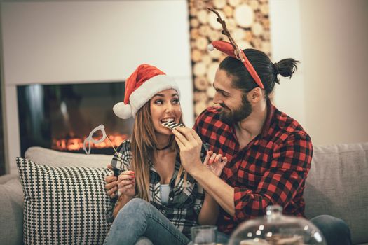 Cute beautiful couple is spending New Year eve at home sitting on sofa and eating cookies. They have great holidays having fun in nice company indoors.