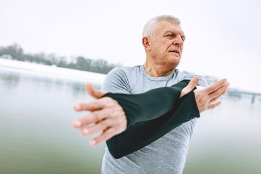 Active senior man stretching and doing exercises by the river during the winter training outside in.