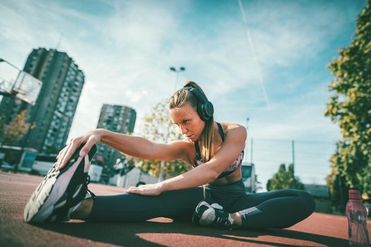 Young female runner, with earphones on her ears, doing stretching exercise on a public place, preparing for morning workout.