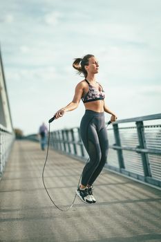 Young female runner jumping over skipping rope and doing exercise on a river bridge, preparing for morning workout.